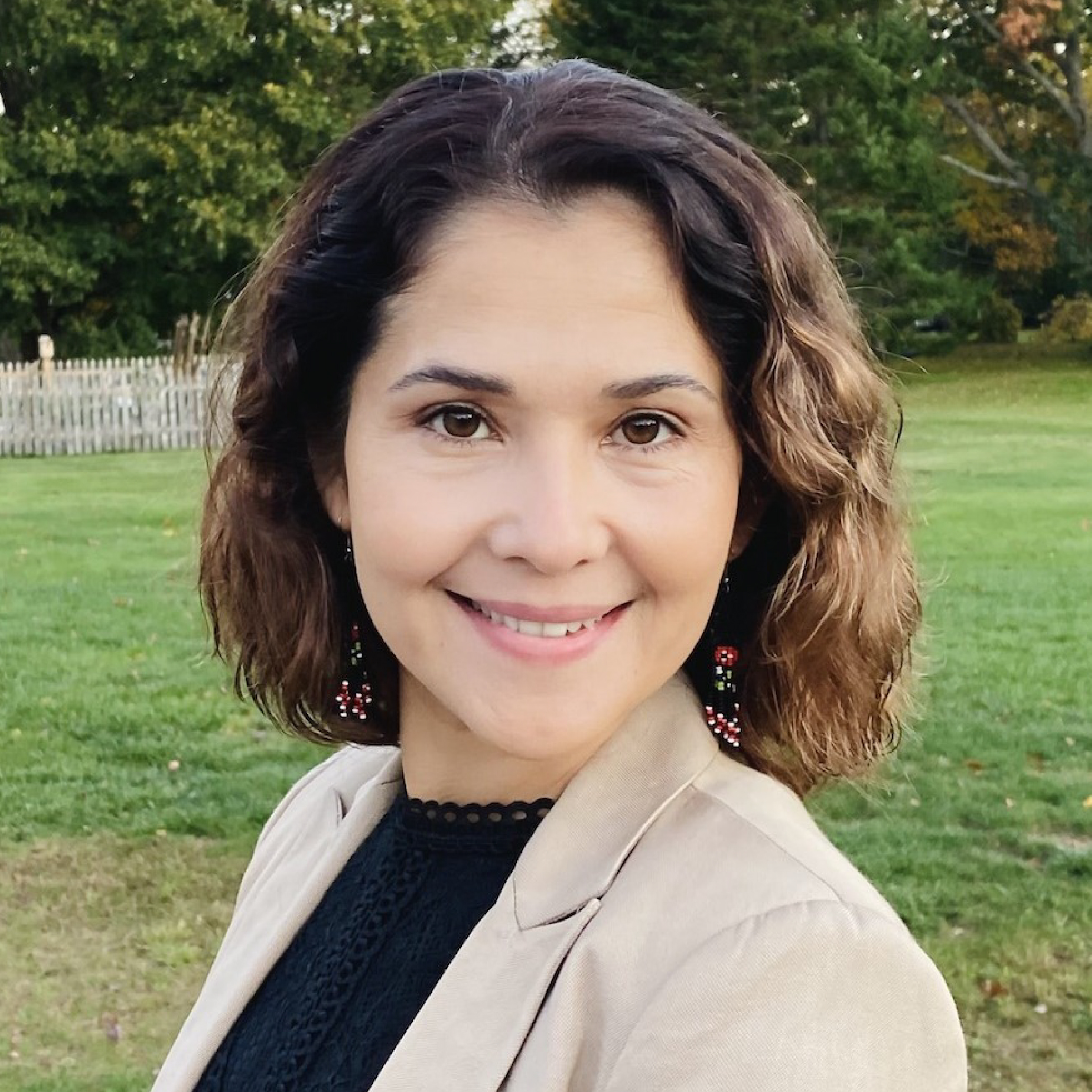 Tavus Begenjova. Woman with wavy brown hair smiling outdoors, wearing a beige jacket and black top, with trees in the background.