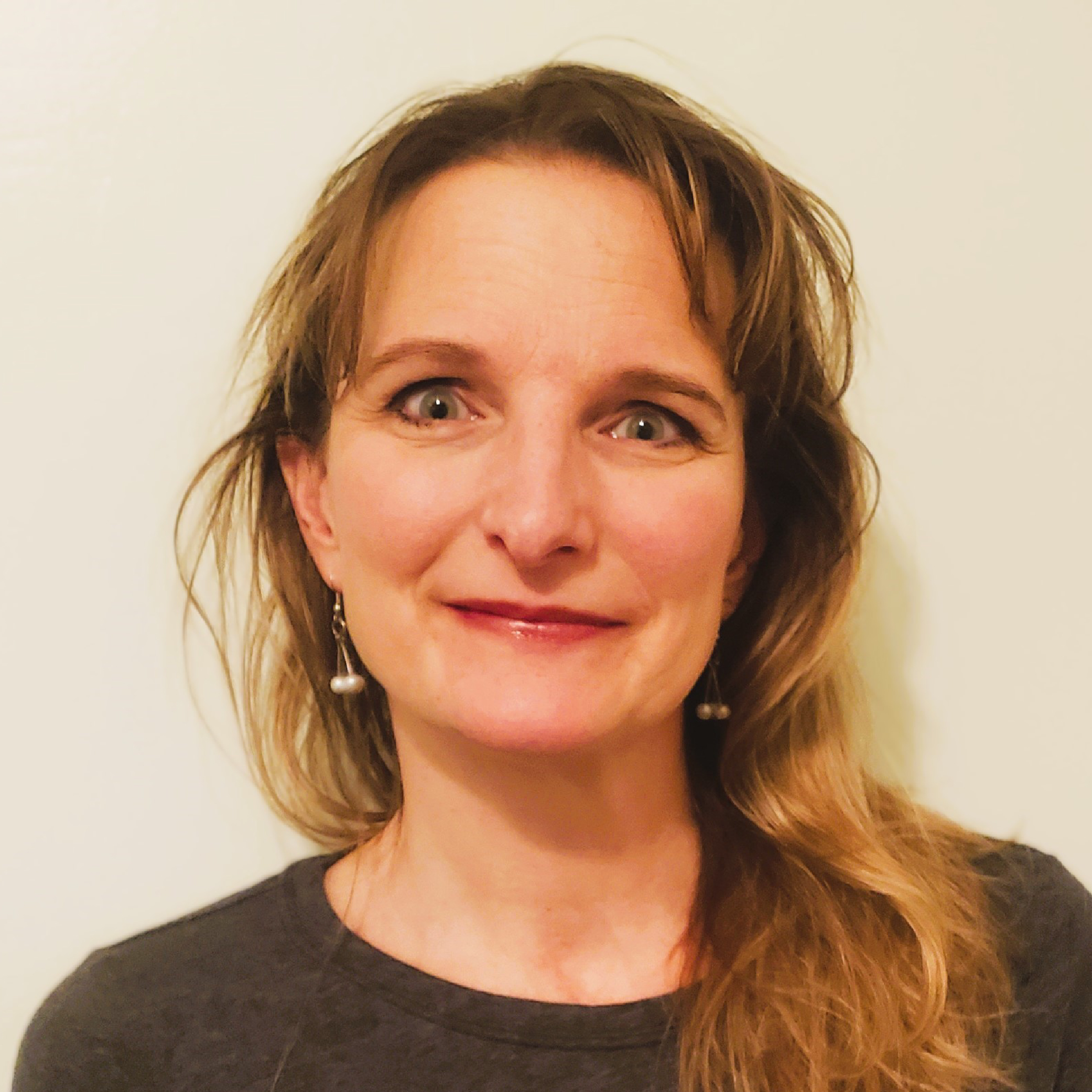 Amy O'Brien. Woman with long wavy blonde hair, wearing earrings and a dark top, smiling in front of a light background.