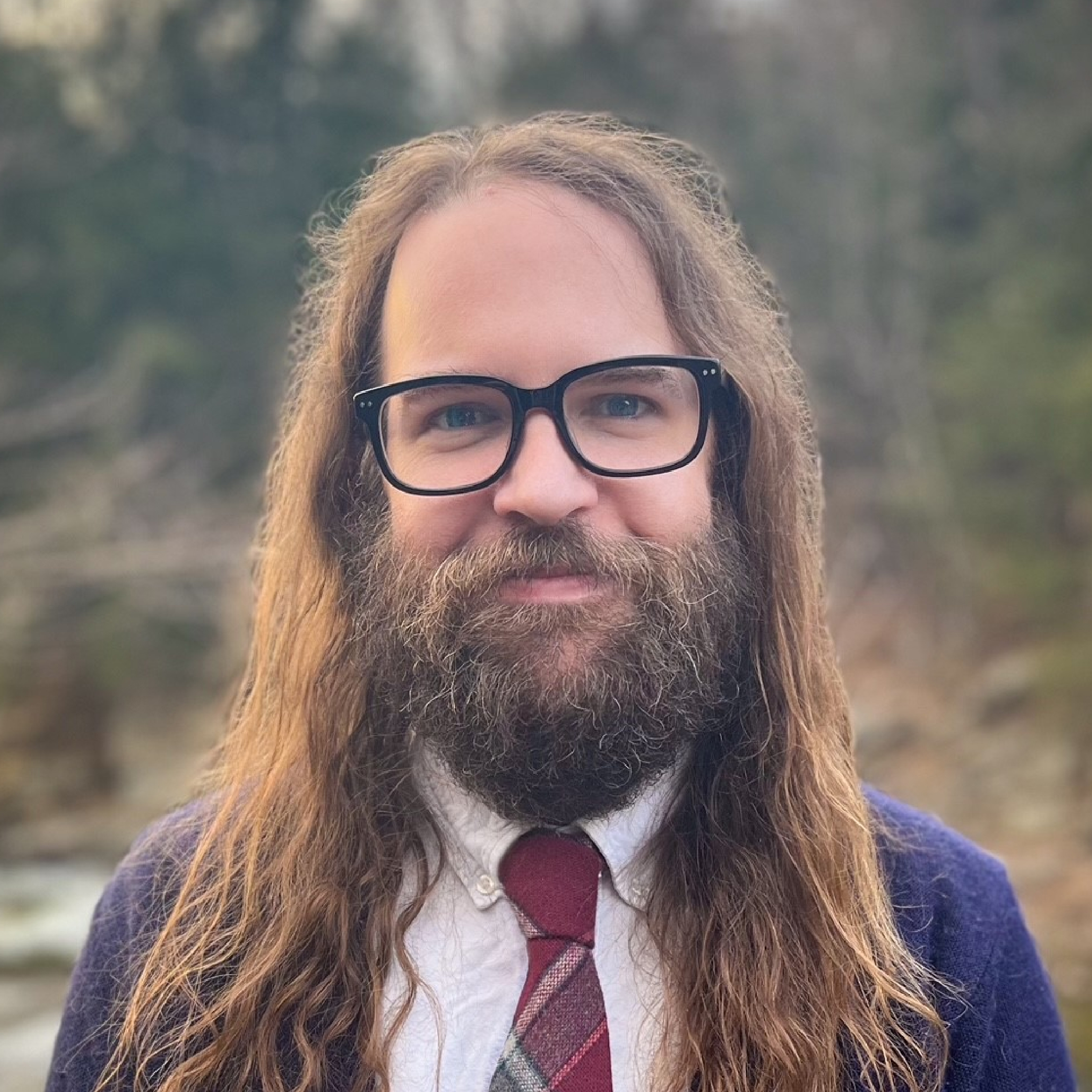 A man with long hair, glasses, and a beard, wearing a tie and sweater, stands outdoors. Danny Kamps