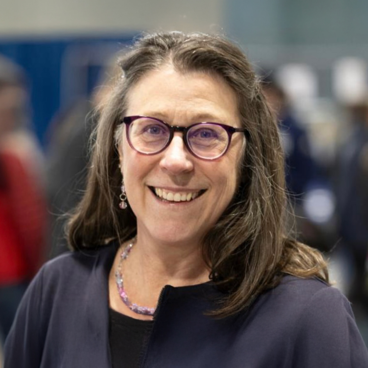 A woman with glasses and long brown hair smiling, wearing a dark top and beaded necklace at an indoor event.