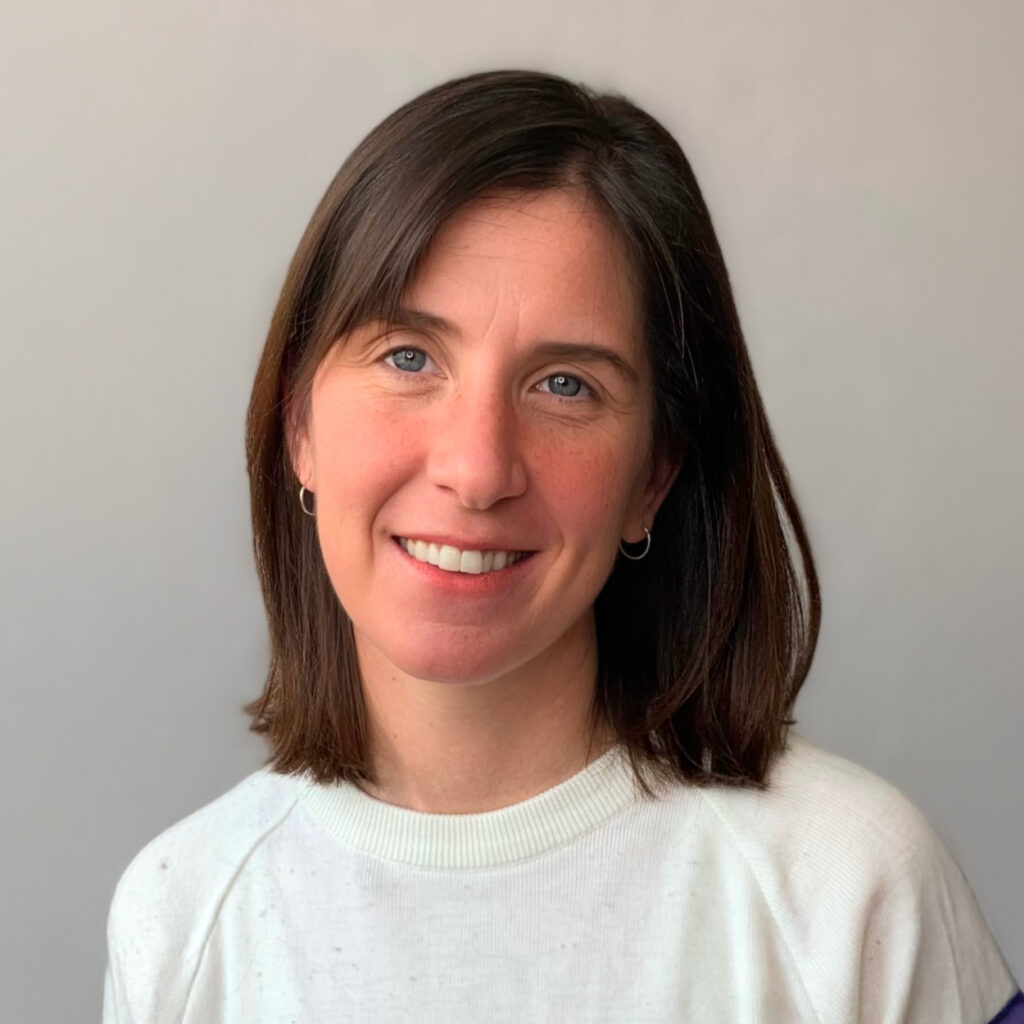 Woman with shoulder-length brown hair, wearing a white sweater, smiling in front of a plain light background.