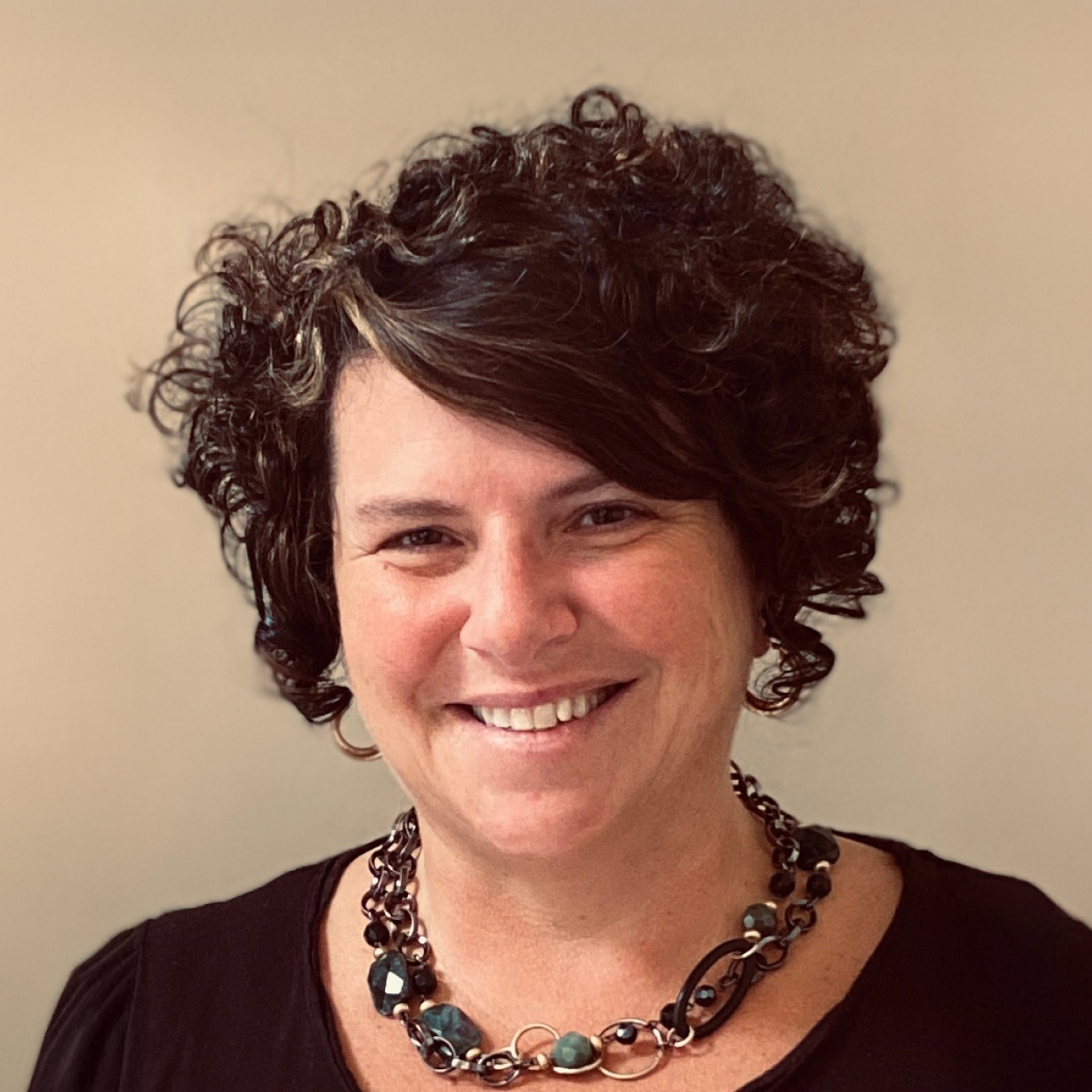 Woman with curly dark hair, smiling, wearing a black top and chunky beaded necklace, on a plain background.
