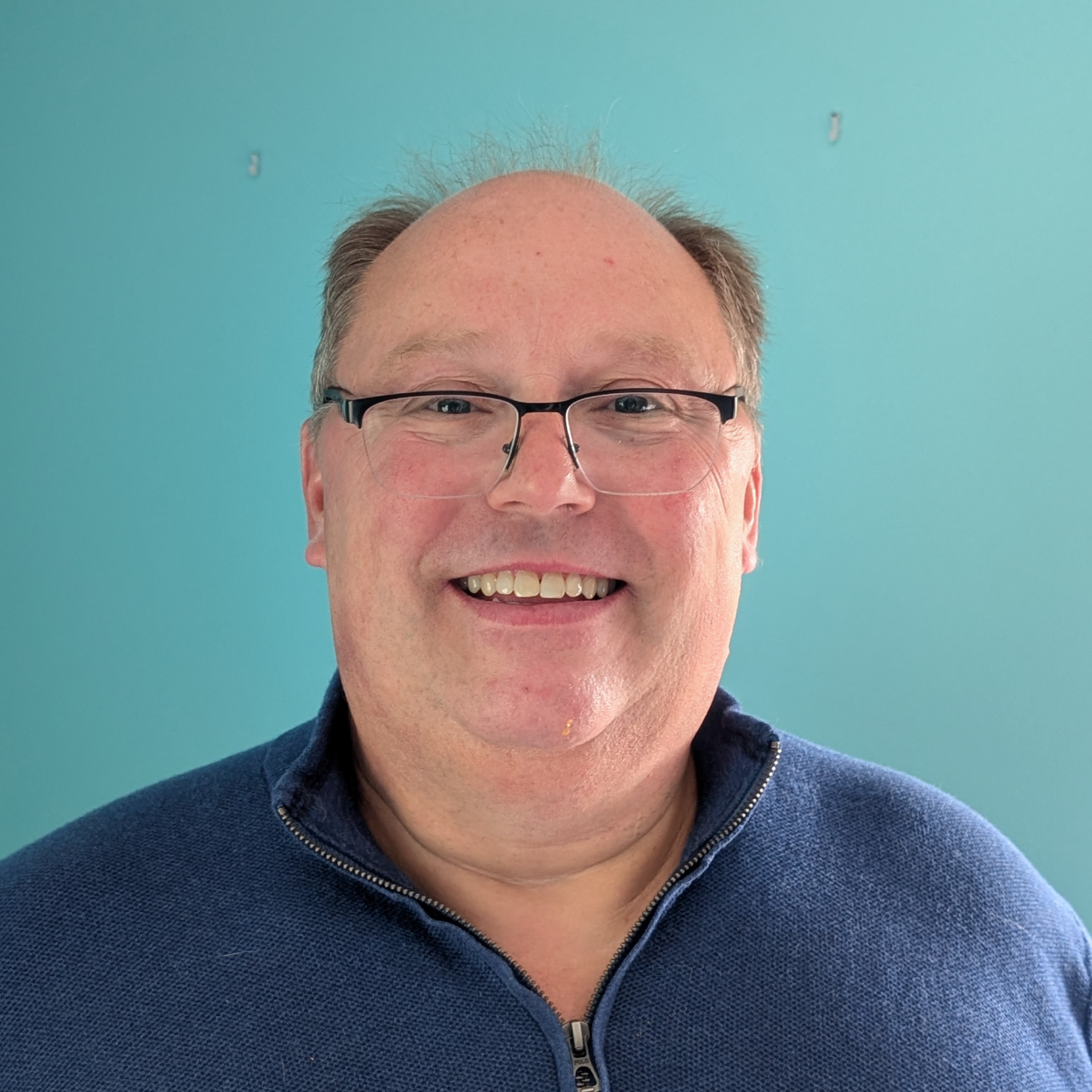 Smiling man with glasses and a blue zip-up sweater in front of a bright turquoise wall.