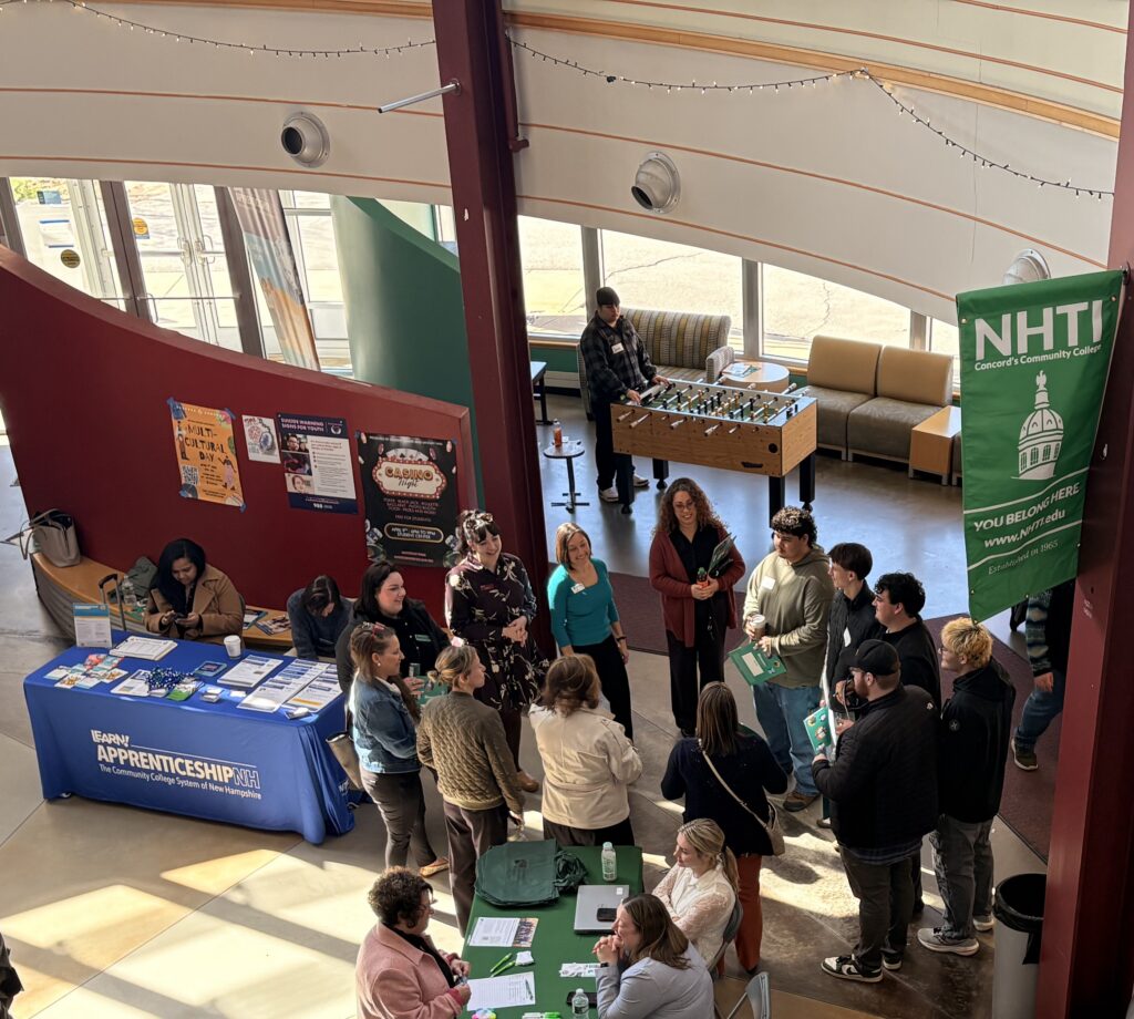 People gather at an indoor career fair with info tables, banners, and brochures in a sunlit space.