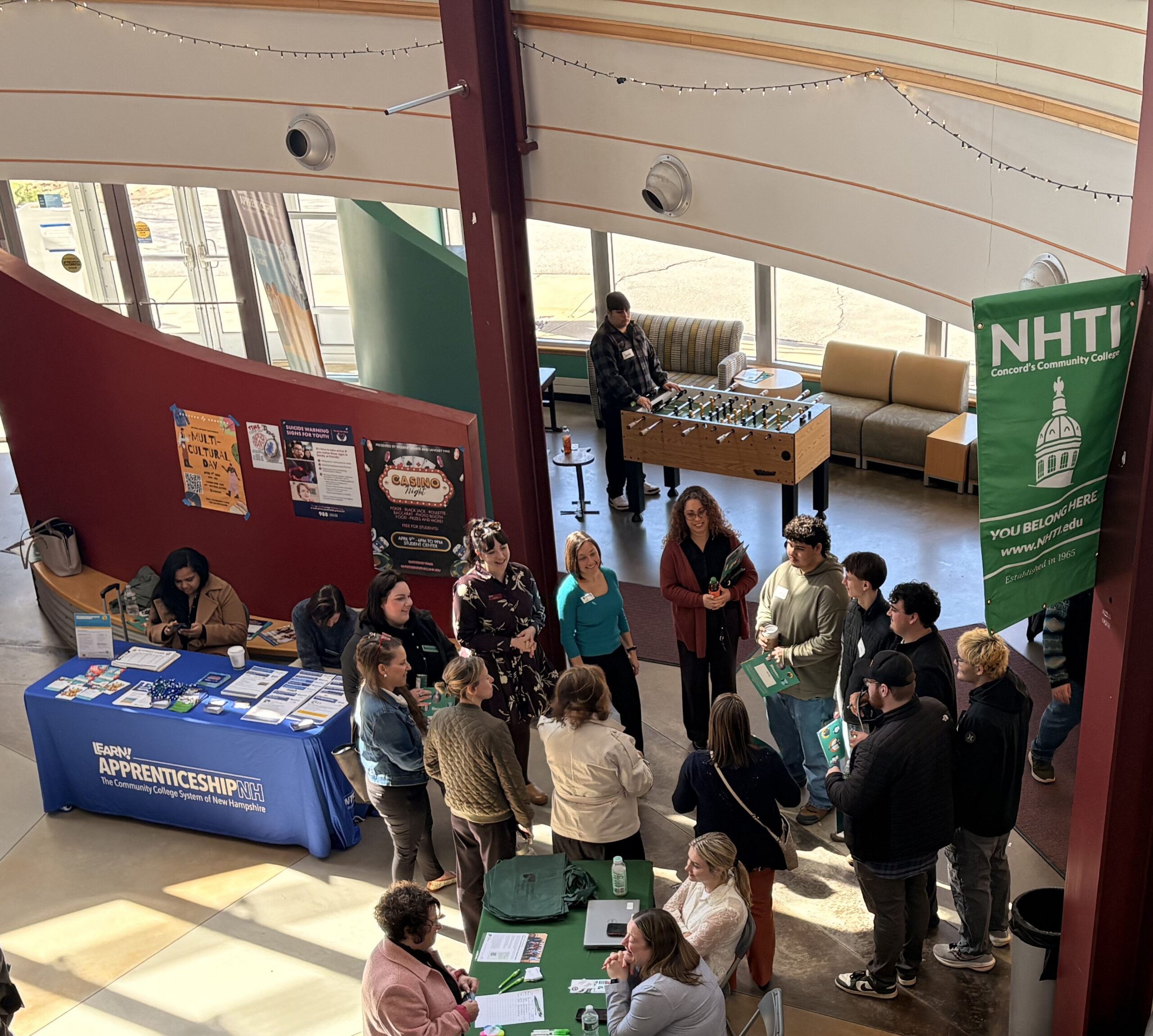 People gather at an indoor career fair with info tables, banners, and brochures in a sunlit space.
