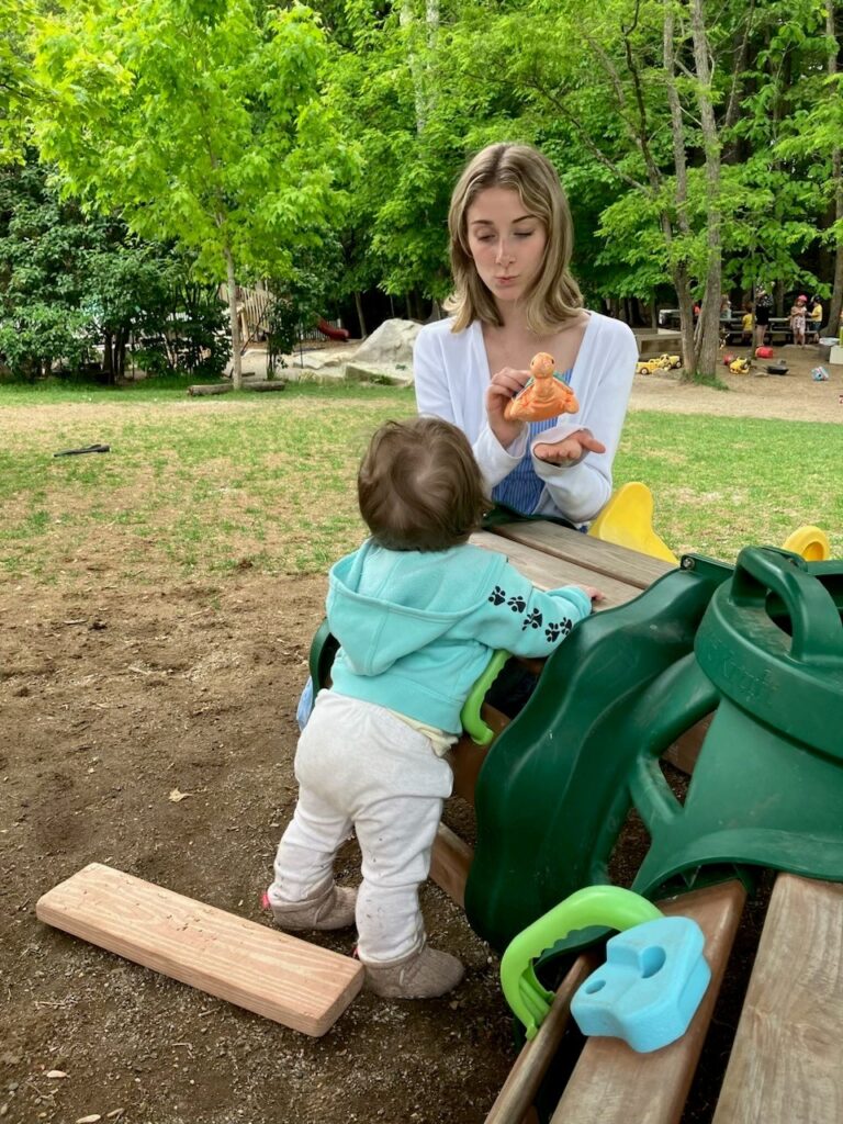 Woman shows a baby a toy while sitting on a bench in a park with green trees in the background.