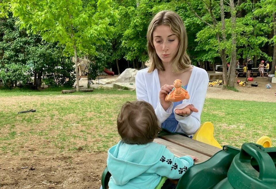 Woman shows a baby a toy while sitting on a bench in a park with green trees in the background.