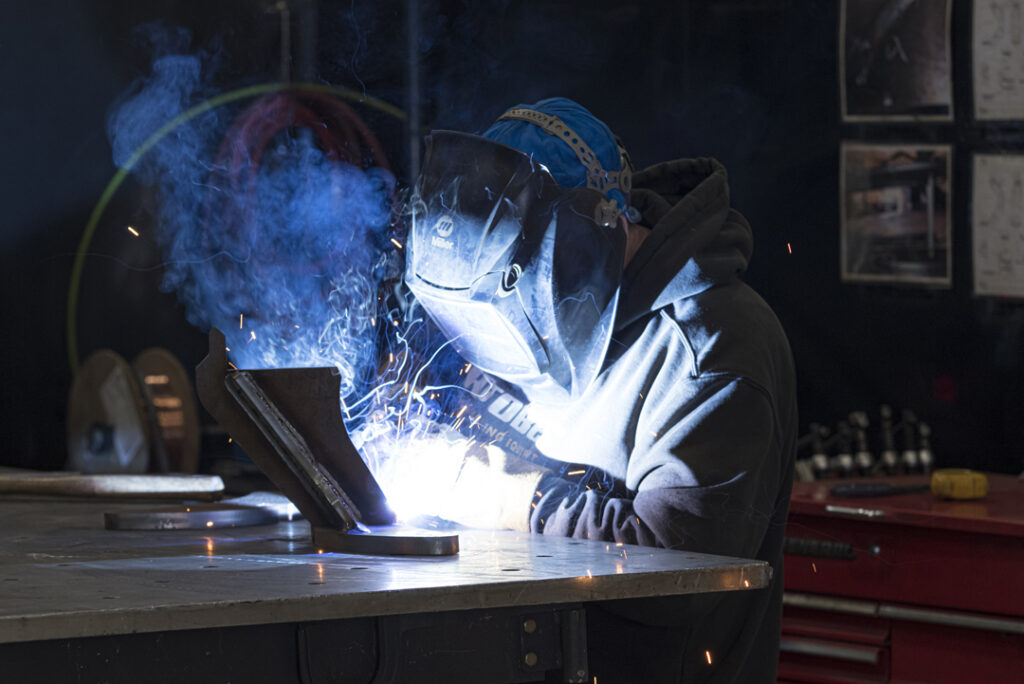 Person welding metal pieces, wearing protective gear, with bright sparks and blue smoke visible.