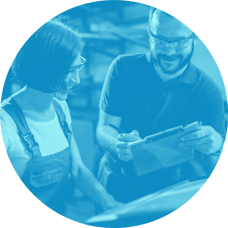 Two people wearing safety glasses smiling and looking at a clipboard in an industrial setting.