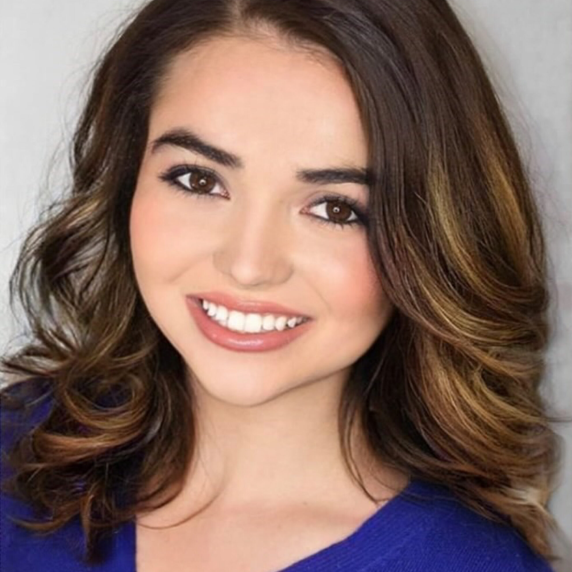 Smiling woman with wavy brown hair, wearing a blue top, posing in front of a light background. Brianna Hayward