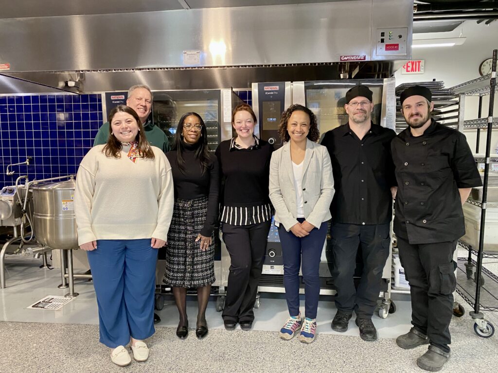 Seven people stand and smile in a commercial kitchen with ovens, blue tiles, and metal shelves behind them.