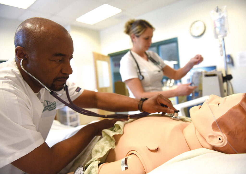 A nurse uses a stethoscope on a medical mannequin while another nurse works in the background.