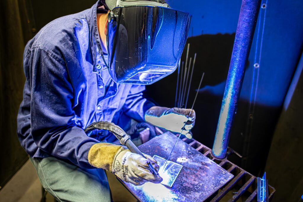 Person wearing gloves and welding helmet working with metal and welding tools under blue lighting.
