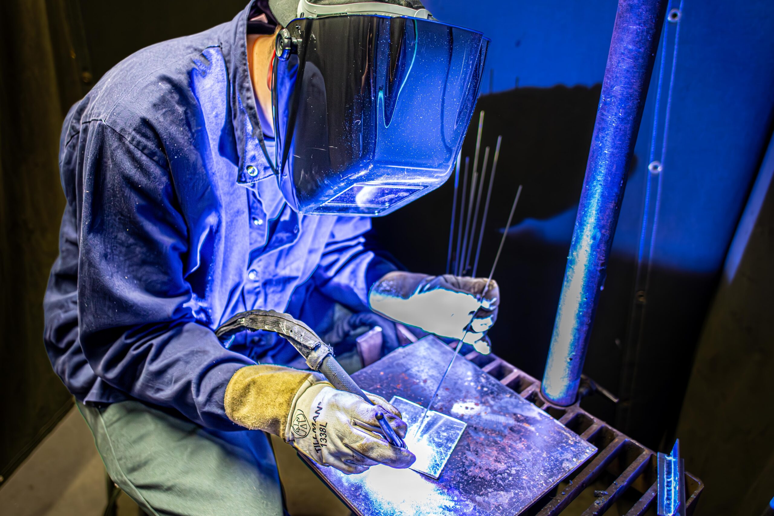 Person wearing gloves and welding helmet working with metal and welding tools under blue lighting.