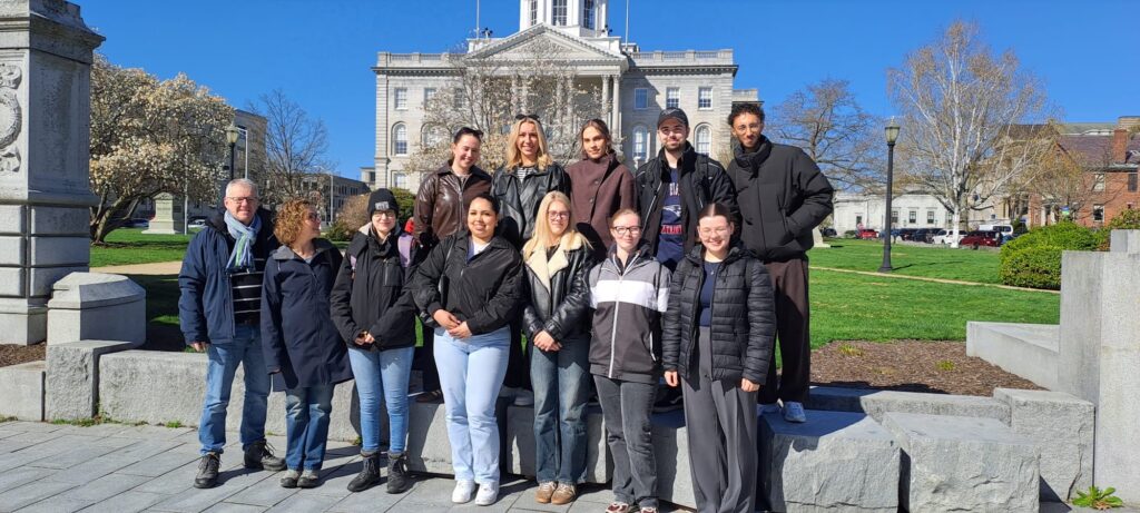 A group of people pose together outside in front of a large historic building on a sunny day.
